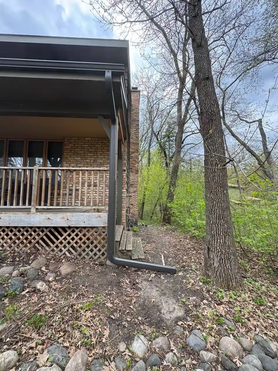 Dark custom downspout with an extension protecting a wooden deck and home foundation on a wooded lot in Eden Prairie.